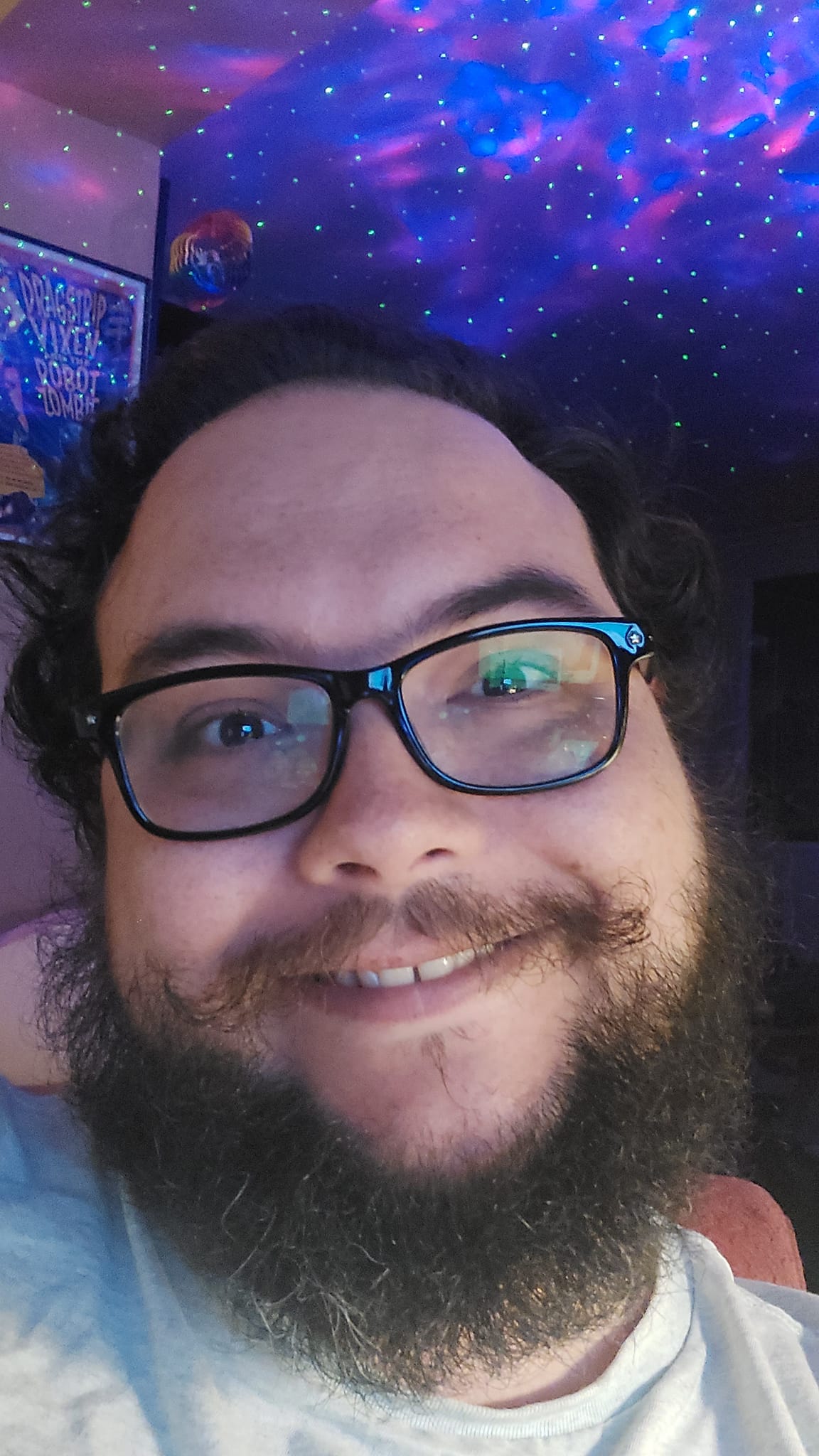 A man with short hair and bushy facial hair sits in his apartment, lights in the background imitate a nebula as captured by the Hubbel Space Telescope